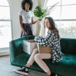 Two young women moving into a bright apartment, sitting on a green sofa with boxes.